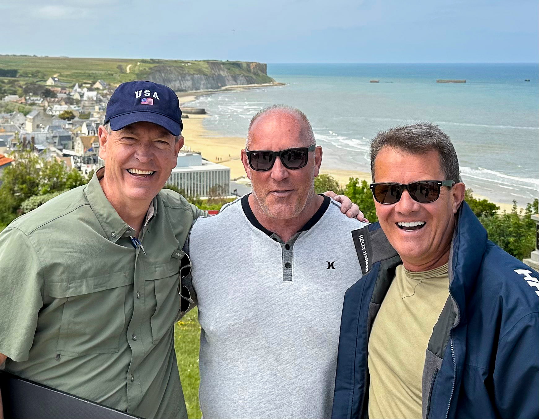 Three travelers above Arromanches, with WWII Mulberry harbor remains in the sea behind