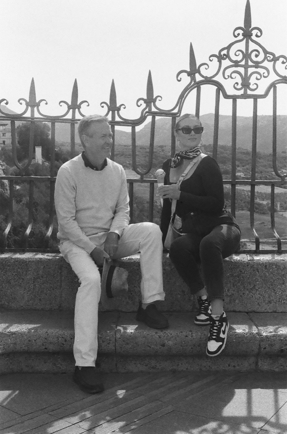 Two travelers resting on a wall beside a wrought-iron railing, shot on black and white film